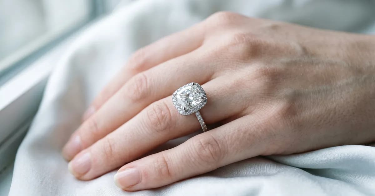 淡い色の布の上に置かれた手、クッションカットのダイヤモンドを小さなダイヤモンドのハローで囲んだリングを身につけている